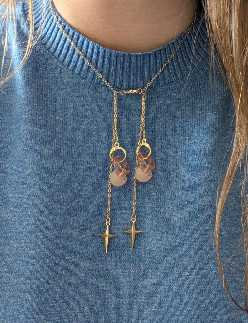 Puede incluir: Un collar tipo lariat en tono dorado con una cadena fina y dos secciones colgantes, cada una adornada con un peque&ntilde;o anillo transl&uacute;cido color melocot&oacute;n, una cuenta esmerilada y un dije de estrella de cuatro puntas en el extremo.