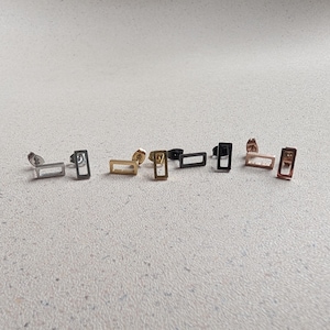 May include: A selection of rectangular stud earrings in various metallic finishes, including silver, gold, black, and rose gold. The earrings have a minimalist, open-frame design, showcasing a modern aesthetic. The earrings are displayed on a light-coloured surface.