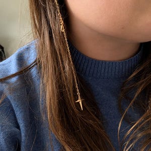 Puede incluir: Un ear cuff dorado conectado a una cadena larga y delicada que cuelga hacia abajo. La cadena presenta un peque&ntilde;o dije de estrella de cuatro puntas en el extremo inferior. El ear cuff est&aacute; colocado en el cart&iacute;lago superior de la oreja.
