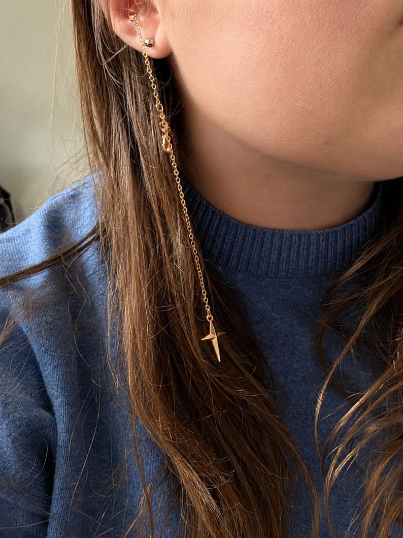 Puede incluir: Un ear cuff dorado conectado a una cadena larga y delicada que cuelga hacia abajo. La cadena presenta un peque&ntilde;o dije de estrella de cuatro puntas en el extremo inferior. El ear cuff est&aacute; colocado en el cart&iacute;lago superior de la oreja.