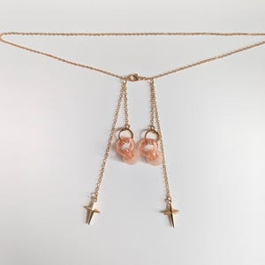 Puede incluir: Un collar de cadena en tono dorado con un cierre central y dos hilos de cadena colgantes. Cada hilo termina en un dije de estrella de cuatro puntas, y la parte superior de cada hilo sostiene un peque&ntilde;o anillo circular transl&uacute;cido color melocot&oacute;n.