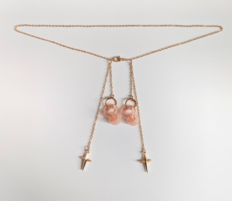 Puede incluir: Un collar de cadena en tono dorado con un cierre central y dos hilos de cadena colgantes. Cada hilo termina en un dije de estrella de cuatro puntas, y la parte superior de cada hilo sostiene un peque&ntilde;o anillo circular transl&uacute;cido color melocot&oacute;n.