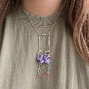 May include: A silver-toned chain necklace featuring two lavender-coloured earplugs attached to the links. The necklace includes multiple small, dangling, multi-coloured beads in shades of pink, blue, green, and orange, suspended from the silver chains.