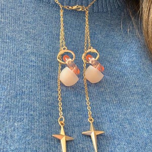 Puede incluir: Un collar tipo lariat en tono dorado con dos cadenas delicadas unidas a un cierre central. Cada cadena est&aacute; adornada con un anillo naranja transl&uacute;cido, una cuenta blanca mate y un dije de estrella de cuatro puntas en cada extremo.