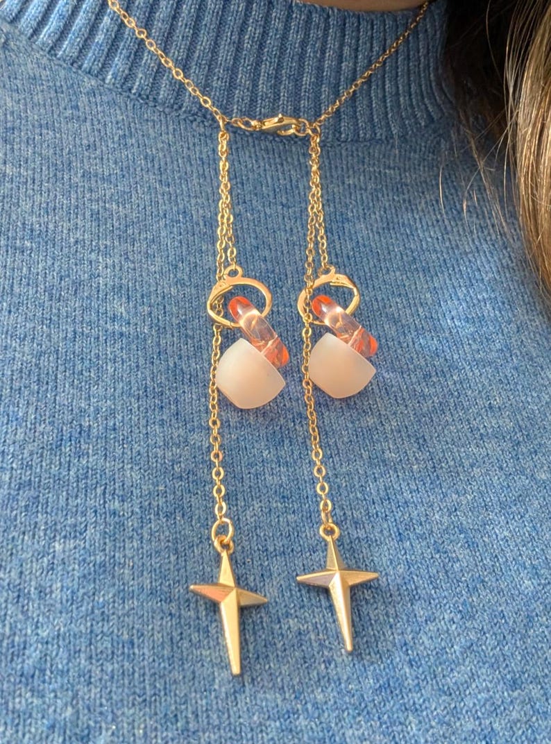 Puede incluir: Un collar tipo lariat en tono dorado con dos cadenas delicadas unidas a un cierre central. Cada cadena est&aacute; adornada con un anillo naranja transl&uacute;cido, una cuenta blanca mate y un dije de estrella de cuatro puntas en cada extremo.