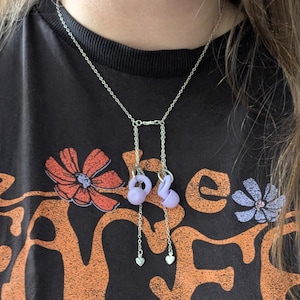 May include: A silver-toned chain necklace featuring two light purple earplugs attached to the main chain by two shorter silver chains, each ending in a small silver heart charm. The necklace is worn over a black t-shirt with orange and purple floral graphics.