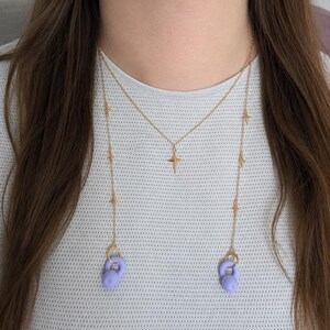 Può includere: Una collana a catena color oro con un ciondolo a croce centrale e piccoli dettagli a forma di stella lungo la catena. Le estremità della catena sono fissate a due accessori arrotondati color lilla chiaro con piccoli anelli color oro.