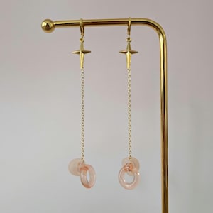 May include: A pair of gold-toned drop earrings featuring a four-pointed star charm attached to a delicate chain. At the base of each chain hangs a translucent, pale pink circular ring component, creating a long, dangling silhouette.