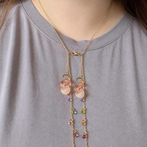 Könnte beinhalten: Eine goldfarbene Kettenhalskette im Lariat-Design mit zwei herabhängenden Strängen. Die Halskette ist mit durchscheinenden, rosafarbenen, muschelartigen Anhängern und kleinen, bunten Perlen in Violett, Grün, Orange und Pink verziert.