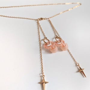 Puede incluir: Un collar de tono dorado con una cadena fina y dos dijes colgantes. Cada dije consiste en una cuenta redondeada, transl&uacute;cida y de color rosa p&aacute;lido unida a un peque&ntilde;o anillo dorado, rematado con un colgante de metal en forma de estrella.