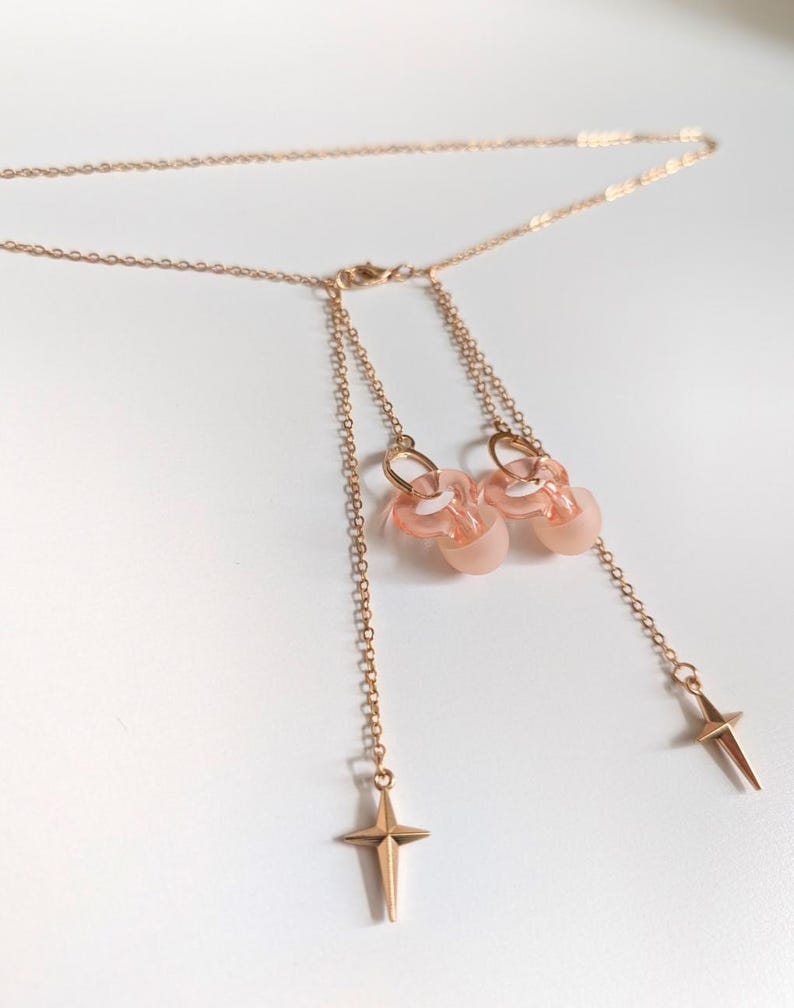 Puede incluir: Un collar de tono dorado con una cadena fina y dos dijes colgantes. Cada dije consiste en una cuenta redondeada, transl&uacute;cida y de color rosa p&aacute;lido unida a un peque&ntilde;o anillo dorado, rematado con un colgante de metal en forma de estrella.