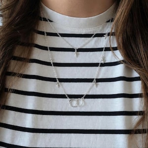 May include: A layered silver-toned necklace featuring two delicate chains. The shorter chain has a small central cross charm, while the longer chain is adorned with multiple small cross charms and two interlocking circular rings at the bottom center.