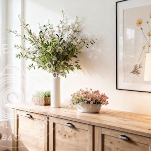 Puede incluir: Un aparador de madera con tiradores negros exhibe un jarrón blanco con vegetación, un pequeño arreglo floral rosa y una impresión botánica enmarcada. Una lámpara y plantas en macetas están a la derecha. Una ventana y un sillón están en el fondo.