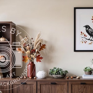 May include: A framed wall art print featuring a black crow perched on a branch with autumn-coloured leaves and berries. The artwork is displayed in a black frame on a light-coloured wall above a wooden cabinet with various decorative items.