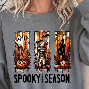 May include: Gray sweatshirt featuring a Halloween-themed graphic. The design includes a ghost, scarecrow, and haunted house, with pumpkins and the text "Spooky Season". The artwork is in orange, black, and white.