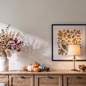 May include: A wooden sideboard features a white vase with dried autumn foliage, a framed botanical print of pumpkins and leaves, a table lamp with a white shade, a bowl of nuts, a stack of books, and a collection of small decorative pumpkins.