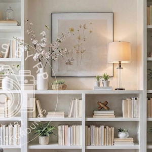 May include: White bookshelves filled with books, plants, and decorative items. A framed botanical print hangs above a lamp with a clear glass base. The shelves are arranged in a symmetrical design, creating a clean and organized aesthetic.