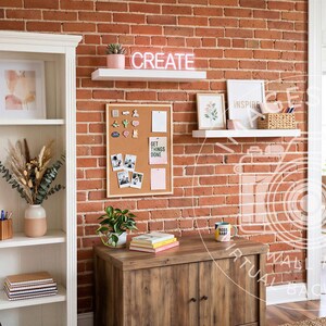 Op de afbeelding: Een witte boekenkast gevuld met kleurrijke boeken, een plant in pot en droogbloemen. Een bakstenen muur bevat een prikbord, twee witte zwevende planken met een neon CREATE-bord, ingelijste kunst en een houten kast met een plant en notitieblokken.