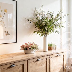 Puede incluir: Escena interior bien iluminada con un mueble de madera, una lámpara y obras de arte enmarcadas. Un jarrón blanco alto contiene vegetación, y una pequeña planta en maceta está cerca. Se ven una ventana y un sillón gris.