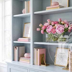 May include: A light blue bookshelf filled with books in various colors, including pink and beige. A vase of pink flowers sits on a shelf, and framed artwork and decorative objects are also present. The bookshelf is positioned near a window.
