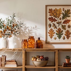 May include: A wooden console table displays a framed botanical print of oak leaves and acorns, a vase with autumn foliage, amber glass jars with fairy lights, a bowl of decorative gourds, a table lamp, and stacked books on its shelves.