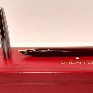 Puede incluir: Una pluma estilogr&aacute;fica burdeos con plum&iacute;n dorado y capuch&oacute;n plateado, sobre una caja roja con letras doradas. Un instrumento de escritura cl&aacute;sico, perfecto para caligraf&iacute;a o uso diario.