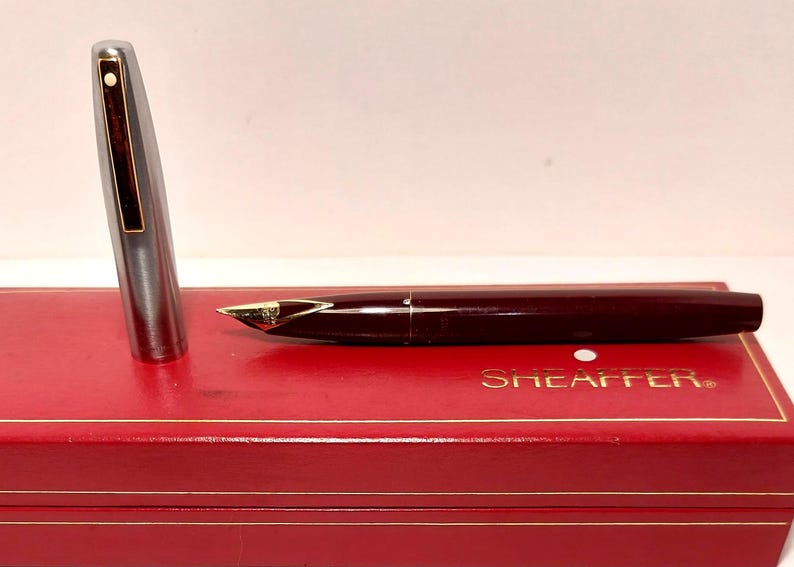 Puede incluir: Una pluma estilogr&aacute;fica burdeos con plum&iacute;n dorado y capuch&oacute;n plateado, sobre una caja roja con letras doradas. Un instrumento de escritura cl&aacute;sico, perfecto para caligraf&iacute;a o uso diario.