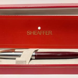 Puede incluir: Un bol&iacute;grafo Sheaffer burdeos y plateado en una caja de presentaci&oacute;n roja. El bol&iacute;grafo tiene un clip dorado y un cuerpo burdeos. La caja es roja con interior blanco y una tarjeta roja con la palabra "SHEAFFER" en letras doradas.