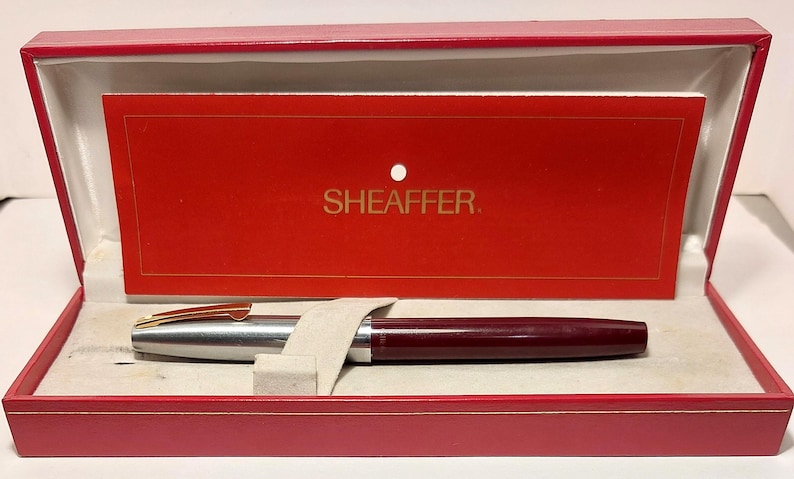 Puede incluir: Un bol&iacute;grafo Sheaffer burdeos y plateado en una caja de presentaci&oacute;n roja. El bol&iacute;grafo tiene un clip dorado y un cuerpo burdeos. La caja es roja con interior blanco y una tarjeta roja con la palabra "SHEAFFER" en letras doradas.