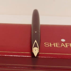 Puede incluir: Una pluma estilogr&aacute;fica burdeos oscuro con plum&iacute;n dorado, sobre una caja roja. El bol&iacute;grafo tiene un dise&ntilde;o elegante. La caja tiene ribetes dorados y la palabra "SHEAFF" es visible.