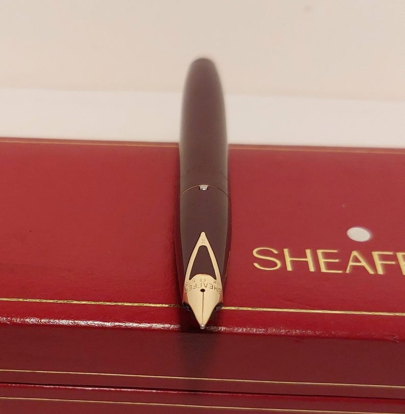 Puede incluir: Una pluma estilogr&aacute;fica burdeos oscuro con plum&iacute;n dorado, sobre una caja roja. El bol&iacute;grafo tiene un dise&ntilde;o elegante. La caja tiene ribetes dorados y la palabra "SHEAFF" es visible.