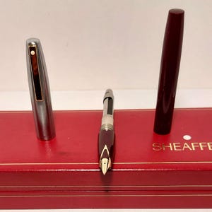 Puede incluir: Un juego de pluma estilogr&aacute;fica Sheaffer vintage con un cuerpo burdeos y una tapa plateada, exhibido en una caja roja. El plum&iacute;n de la pluma es dorado. La tapa de la pluma est&aacute; quitada, revelando el plum&iacute;n y el dep&oacute;sito de tinta.
