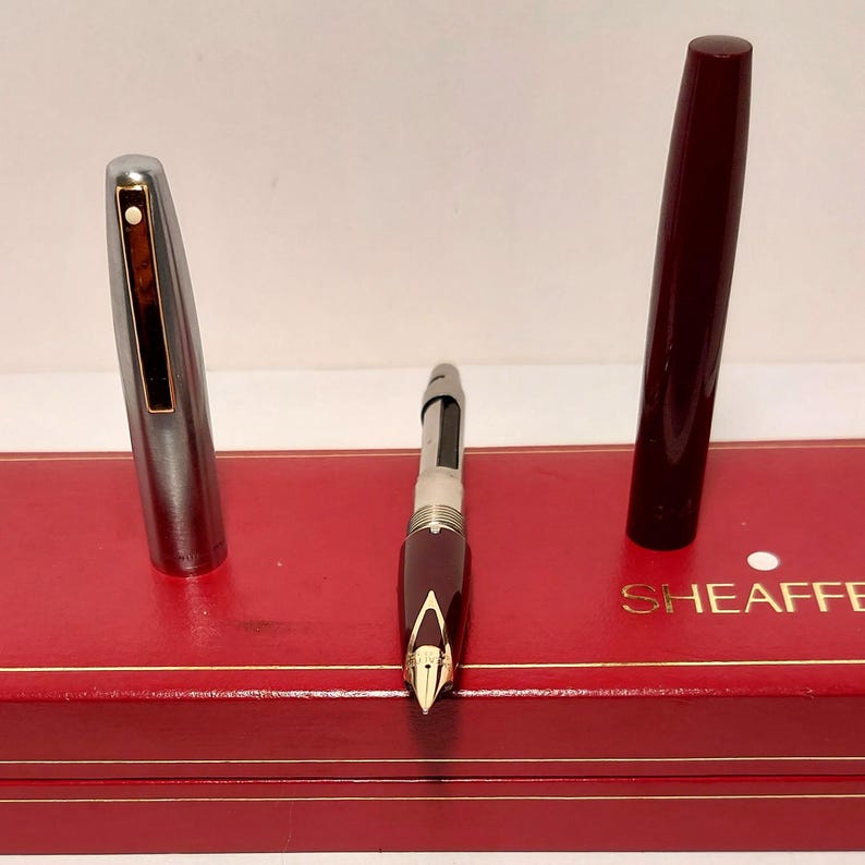 Puede incluir: Un juego de pluma estilogr&aacute;fica Sheaffer vintage con un cuerpo burdeos y una tapa plateada, exhibido en una caja roja. El plum&iacute;n de la pluma es dorado. La tapa de la pluma est&aacute; quitada, revelando el plum&iacute;n y el dep&oacute;sito de tinta.