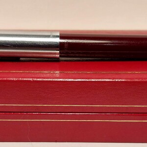 Puede incluir: Una pluma estilogr&aacute;fica burdeos y plateada con un clip dorado descansa sobre una caja roja con ribetes dorados. La pluma se divide en dos secciones, con una parte superior de metal plateado y un cuerpo burdeos. La caja es rectangular y tiene un acabado brillante.