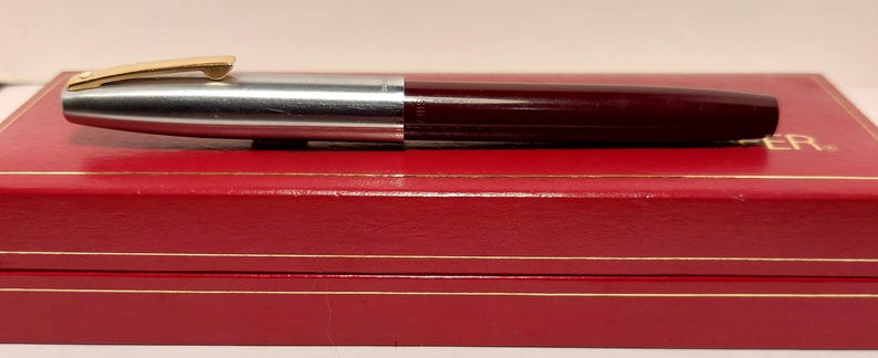 Puede incluir: Una pluma estilogr&aacute;fica burdeos y plateada con un clip dorado descansa sobre una caja roja con ribetes dorados. La pluma se divide en dos secciones, con una parte superior de metal plateado y un cuerpo burdeos. La caja es rectangular y tiene un acabado brillante.