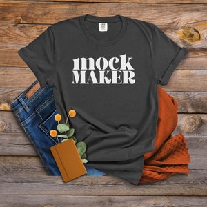May include: A dark gray t-shirt with the text "mock maker" in white letters. The shirt is laid flat on a wooden surface with a pair of blue jeans and a brown notebook.