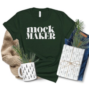 May include: A dark green t-shirt with the text "mock maker" in white letters. The shirt is laid flat on a white background with a brown knitted sweater, a pair of blue jeans, a white mug with a black tree pattern, and a wrapped gift box.