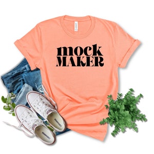 May include: A peach-coloured t-shirt with the words "mock maker" in black, alongside denim shorts and white Converse trainers. Green foliage is arranged around the clothing, creating a casual, lifestyle aesthetic.