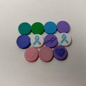 May include: A collection of eleven circular buttons in various colors including teal, green, blue, purple, and pink. Some buttons feature designs such as a light blue and grey awareness ribbon, a cursive letter H, a raised heart, and a textured heart.