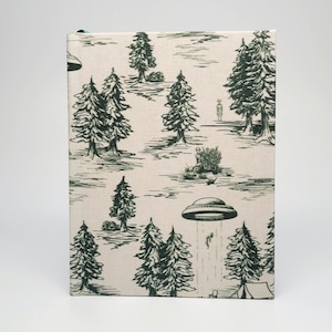 May include: A beige journal with a forest and UFO design. The cover features a repeating pattern of pine trees, flying saucers, and alien figures in a black and white illustration. The journal has a green ribbon bookmark.