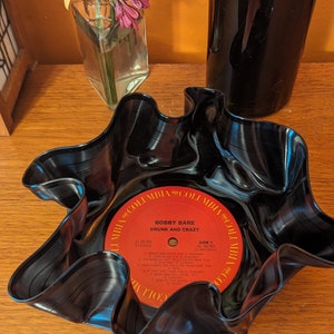 May include: A handcrafted bowl made from a recycled vinyl record. The black record is molded into a wavy, star-like shape, with the original red and white label of "Bobby Bare - Drunk and Crazy" visible in the center. A unique decorative piece.