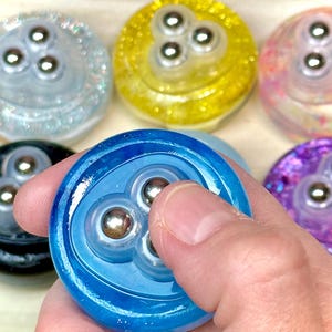 May include: A collection of circular, resin-based fidget tools in various colors including blue, yellow, black, and purple. Each piece features a smooth, translucent surface embedded with three small, polished metal ball bearings arranged in a triangle.
