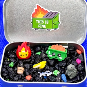 May include: A silver metal tin containing black gravel and several small charms including a red flame, a green dumpster, a coffee cup, a banana, a fish skeleton, and a gray mouse. The inside lid features a sticker of a burning dumpster with the text THIS IS FINE.