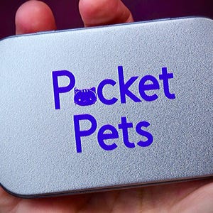 May include: A rectangular, silver-colored tin with the words "Pocket Pets" in bright blue lettering. The word "Pocket" features a cat icon in place of the letter "o". The tin is held in a person's hand.