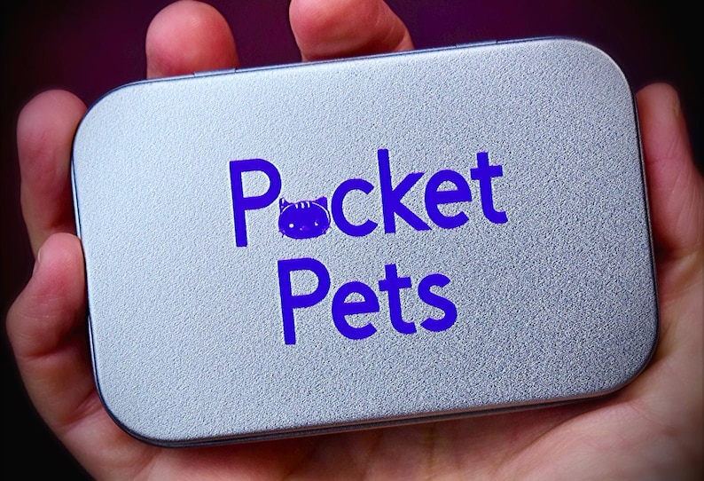 May include: A rectangular, silver-colored tin with the words "Pocket Pets" in bright blue lettering. The word "Pocket" features a cat icon in place of the letter "o". The tin is held in a person's hand.