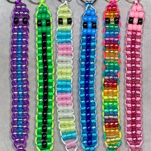 May include: Six beaded keychains featuring a ladder-stitch design with two black beads for eyes at the top. The keychains are made with various colored plastic pony beads, including purple, green, yellow, blue, rainbow, and pink, attached to metal rings.