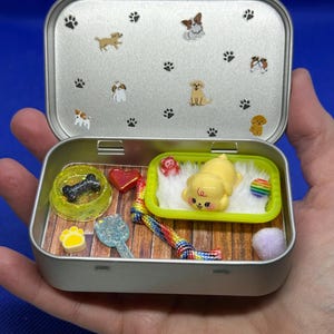 May include: A miniature dog-themed playset inside a silver tin. The tin's lid features paw prints and dog illustrations. Inside, a yellow puppy figure rests on a bed, alongside a bone-shaped treat, a heart, a rainbow toy, and other accessories.
