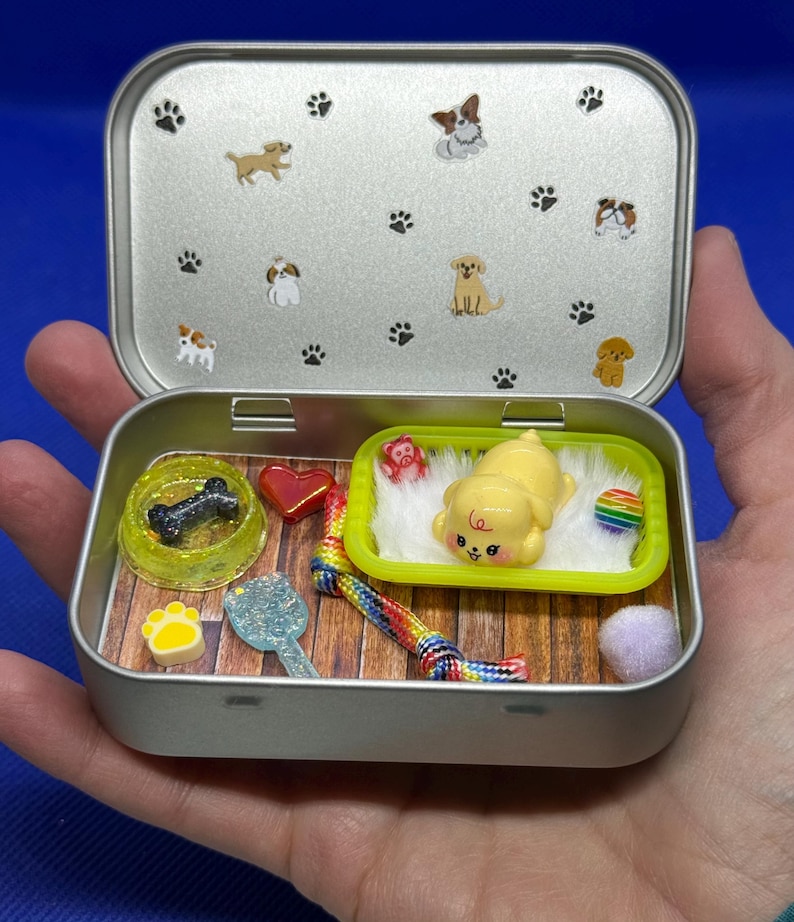 May include: A miniature dog-themed playset inside a silver tin. The tin's lid features paw prints and dog illustrations. Inside, a yellow puppy figure rests on a bed, alongside a bone-shaped treat, a heart, a rainbow toy, and other accessories.
