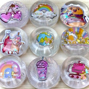 May include: A set of nine circular clear resin cabochons featuring embedded colorful graphics and glitter. Designs include pink cherries, a rainbow with the text DREAM HIGH, a burger, cartoon characters, a globe, a drink, and a pink robot.