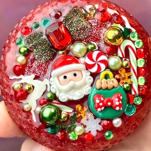 May include: A circular red resin ornament embedded with various Christmas-themed charms including a Santa Claus face, a green bauble with a red polka dot bow, a candy cane, a white reindeer, peppermint candies, and assorted red, green, and gold beads.